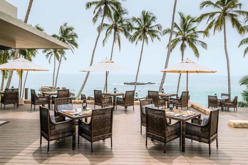 An open dining deck with wicker chairs and cream umbrellas under tall palms, looking out to the sea